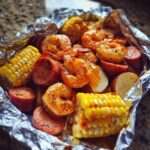 Close-up of cooked shrimp, sausage slices, corn on the cob, and potatoes inside a foil packet, ready for Shrimp Boil Foil Packets.