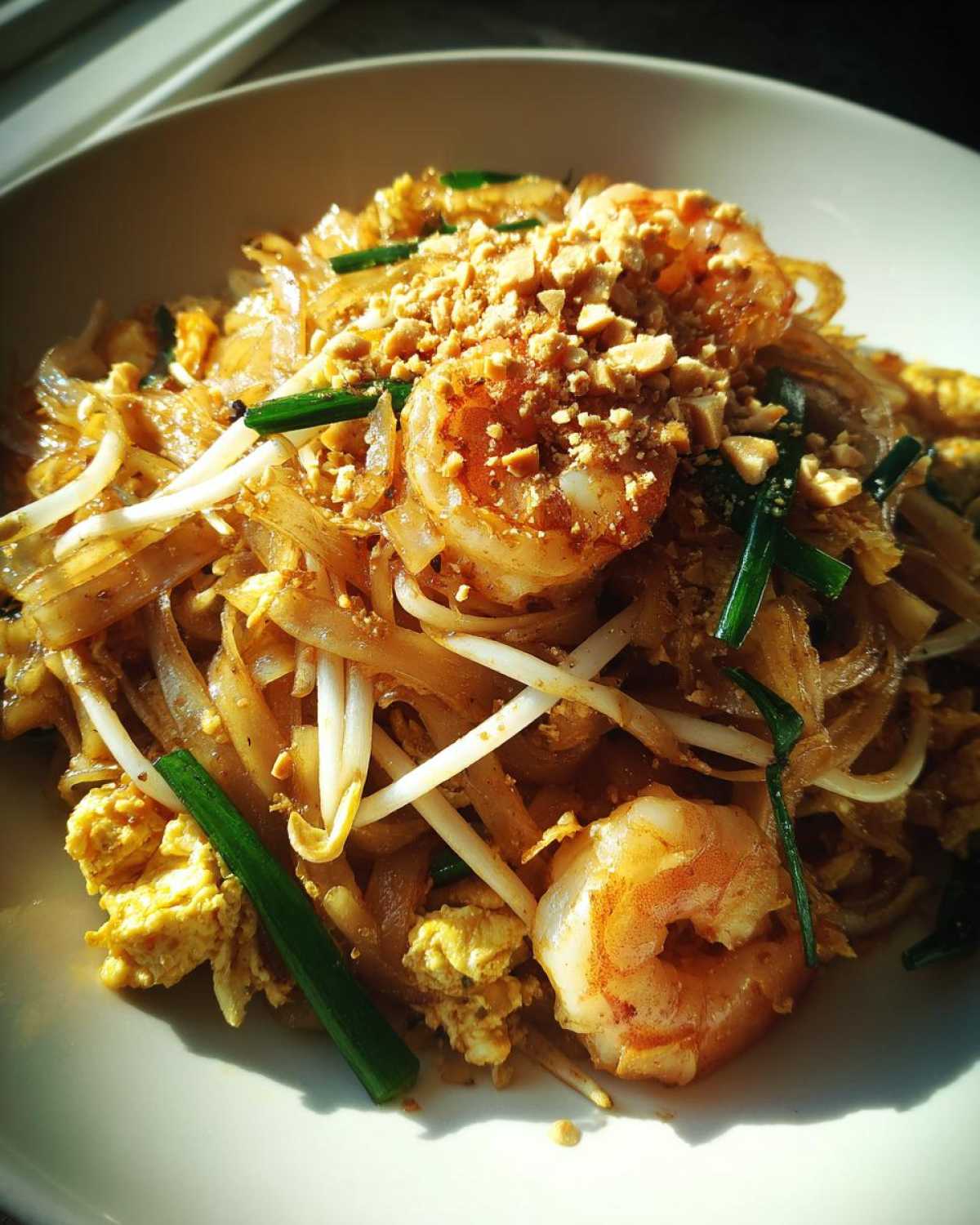 A close-up, brightly lit serving of shrimp Pad Thai with tamarind sauce, topped with crushed peanuts and scallions.