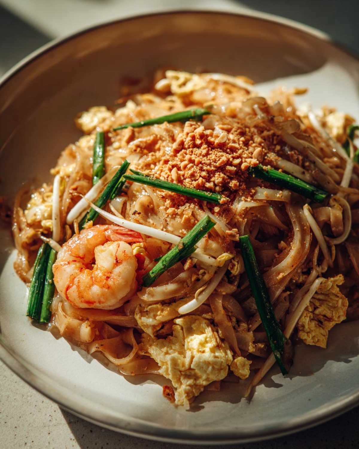 A close-up, sunlit photo of flavorful Pad Thai with Tamarind Sauce, topped with shrimp, crushed peanuts, and green onions.