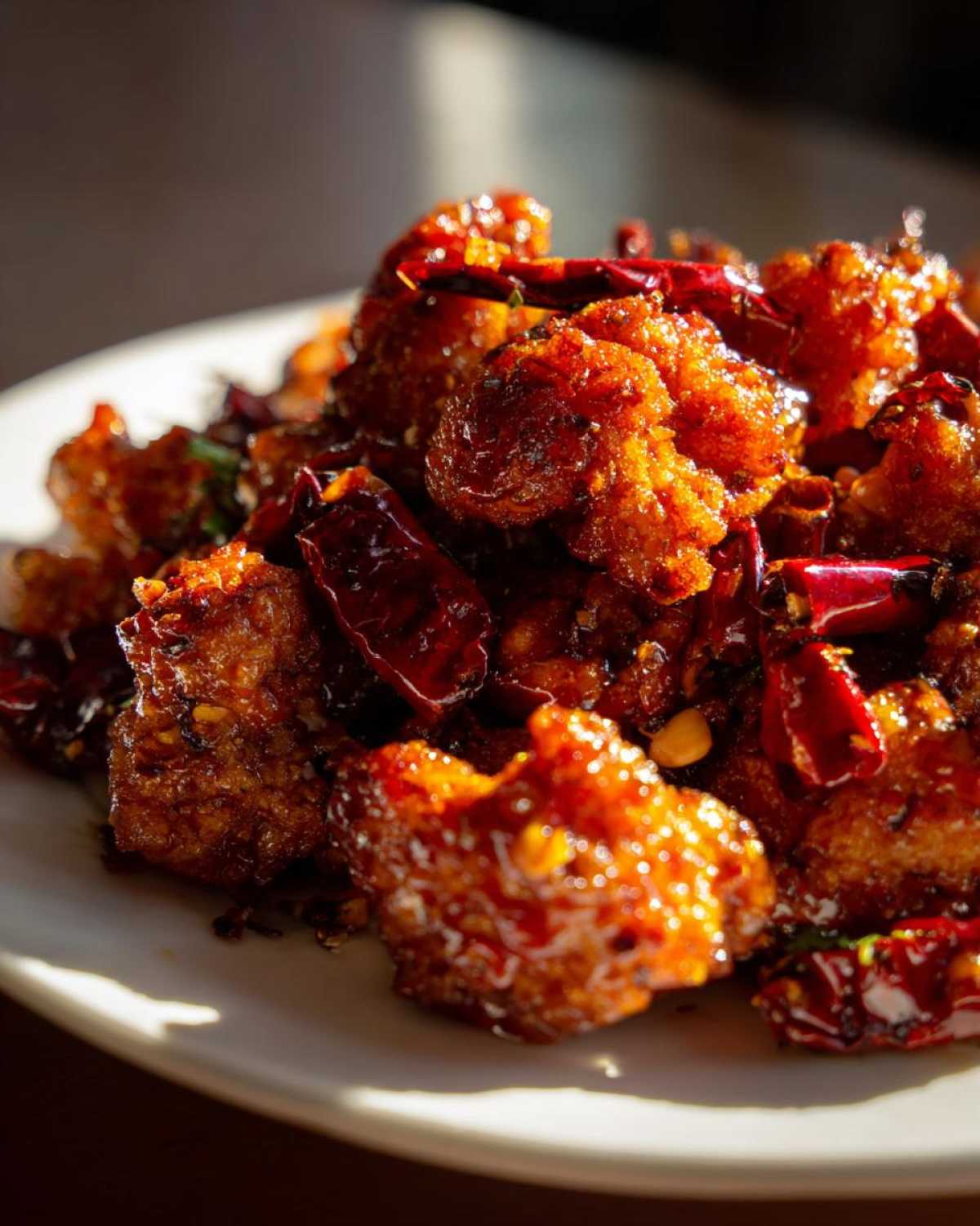 Close-up of crispy, glazed Kung Pao Cauliflower pieces tossed with dried red chilies on a white plate.