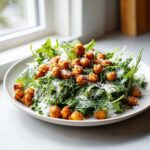A plate of Spring Greens Caesar with Crispy Chickpeas, drizzled with creamy dressing and topped with grated cheese.