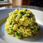 A mound of creamy, bright yellow Spring Pea Mint Risotto studded with vibrant green peas and chopped herbs on a white plate.