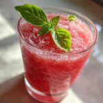 Close-up of a frosty glass filled with bright red Strawberry Basil Lemonade Slush, garnished with fresh basil leaves.