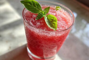 Close-up of a frosty glass filled with bright red Strawberry Basil Lemonade Slush, garnished with fresh basil leaves.