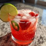 Close-up of a bright red Strawberry Jalapeno Margarita Mocktail filled with ice, strawberries, and a lime wedge garnish.