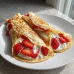 Two rolled Strawberry Mascarpone Crepes filled with white cream and fresh sliced strawberries, served on a white plate.