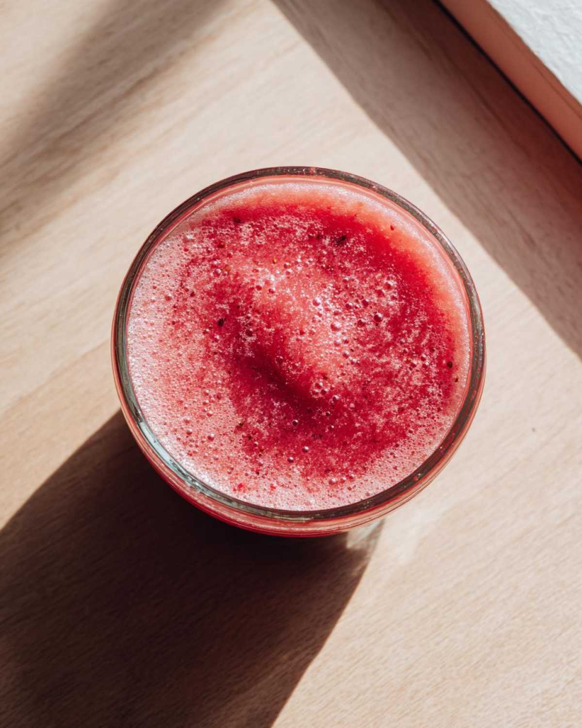 Overhead view of a glass filled with bright red Strawberry Miso Vinaigrette, showing foam on top, sitting on a light wooden surface.