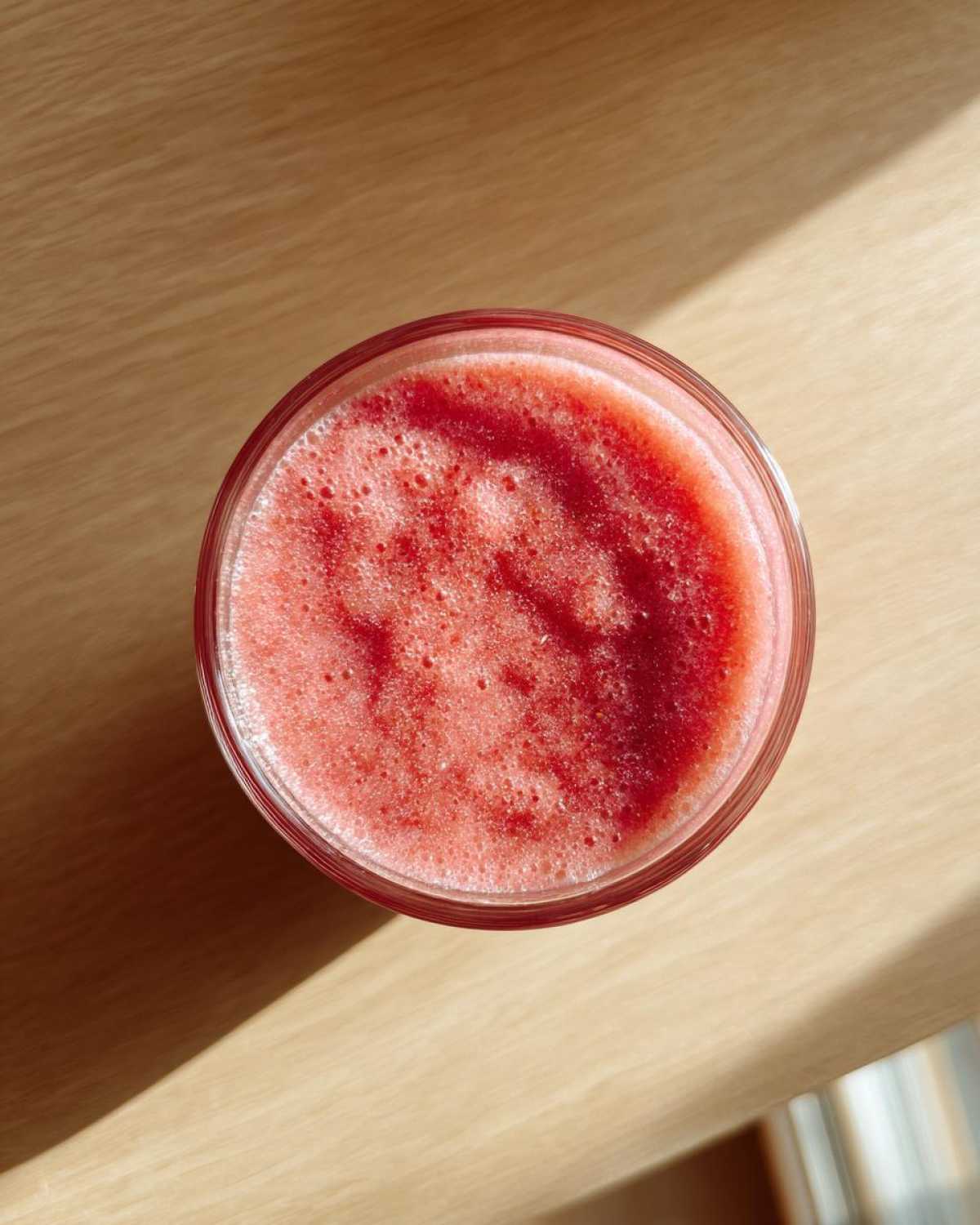 Overhead view of a glass containing a vibrant, frothy red liquid, possibly the Strawberry Miso Vinaigrette, on a light wooden surface.