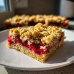 A close-up, sunlit shot of a single Strawberry Oat Crumble Bar slice showing the thick oat base and bright red strawberry filling.