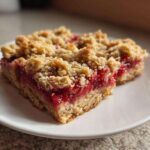 Two squares of rich Strawberry Oat Crumble Bars featuring a thick jam layer and crumbly oat topping, served on a white plate.