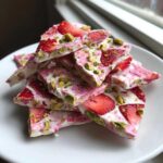 A stack of broken pieces of Strawberry Yogurt Bark with Pistachios featuring sliced strawberries and green nuts on a white plate.