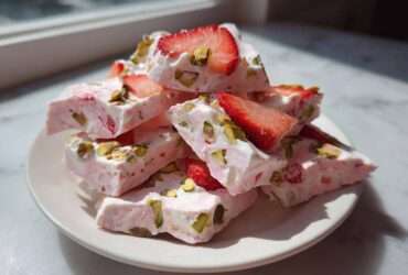 A stack of broken pieces of pink and white Strawberry Yogurt Bark with Pistachios and fresh strawberry slices on a white plate.