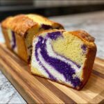 A thick slice of Ube Coconut Swirl Bread showing vibrant purple ube swirls against the yellow bread dough, resting on a wooden cutting board.