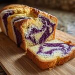 A loaf of Ube Coconut Swirl Bread, partially sliced, showing vibrant purple ube swirls against the yellow and white batter on a wooden board.