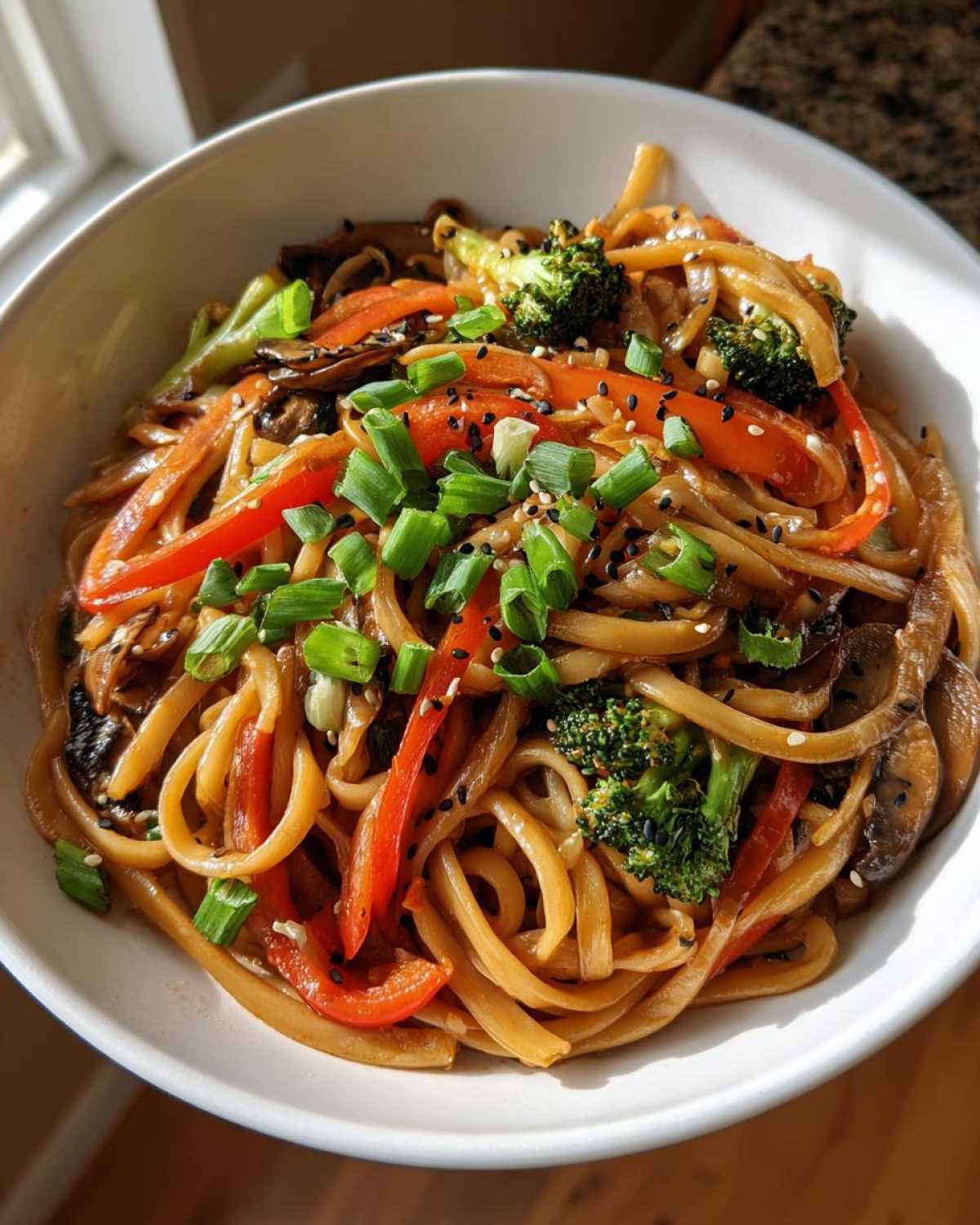 A close-up of a white bowl filled with Udon Stir-Fry with Sesame Vegetables, topped with scallions and sesame seeds.