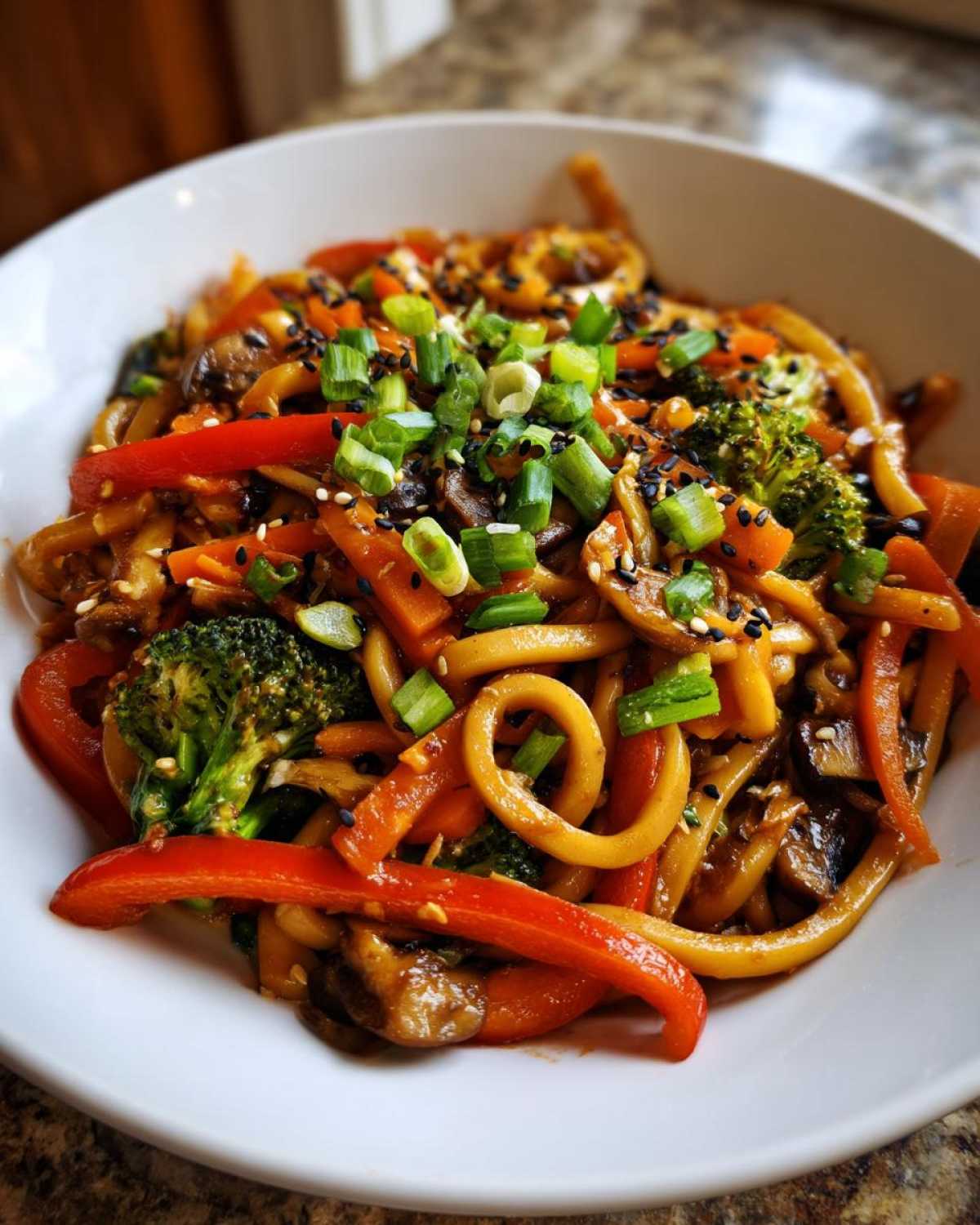 A close-up of a white bowl filled with Udon Stir-Fry with Sesame Vegetables, topped with scallions and black sesame seeds.