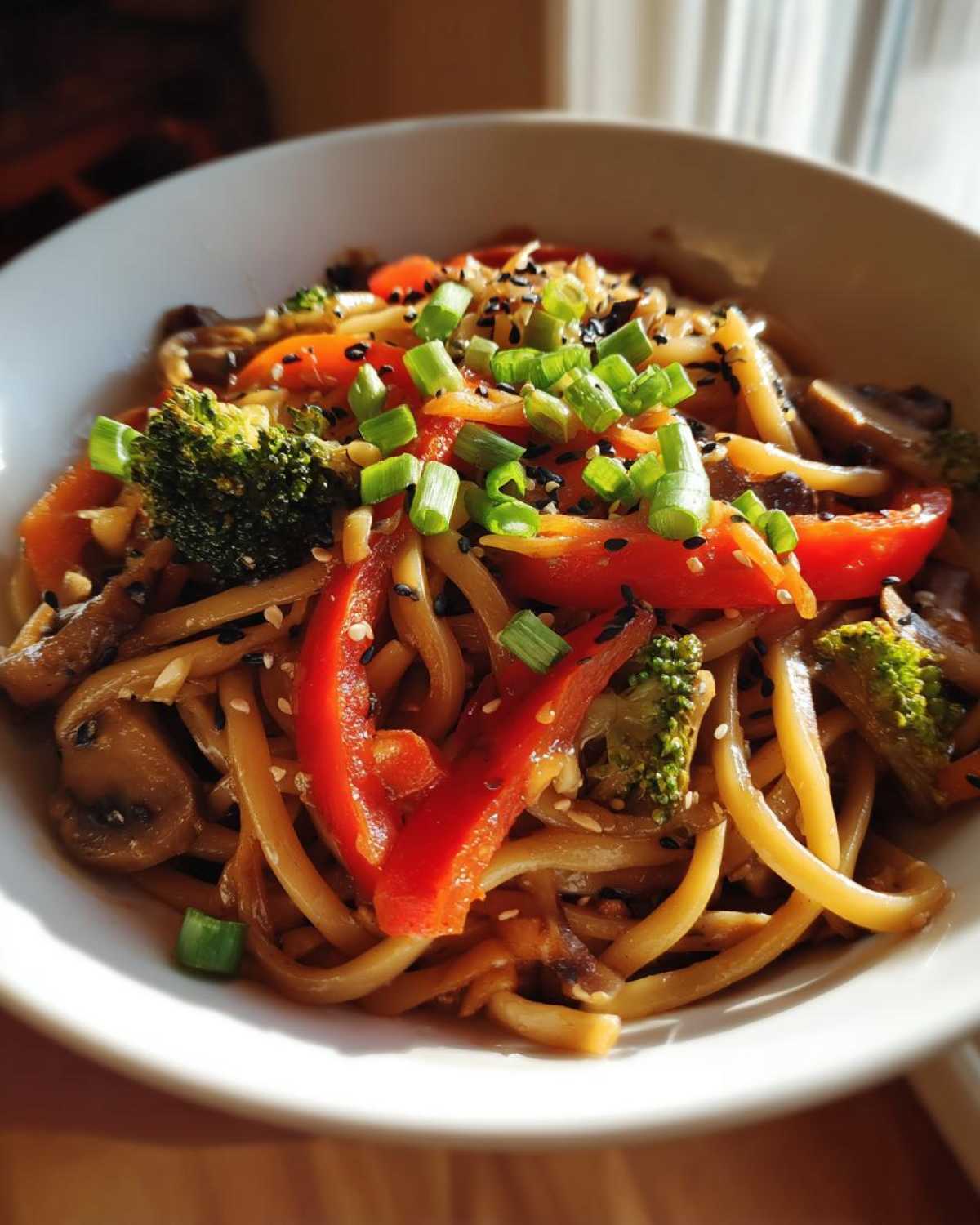 A close-up bowl of Udon Stir-Fry with Sesame Vegetables, topped with red peppers, broccoli, and green onions.