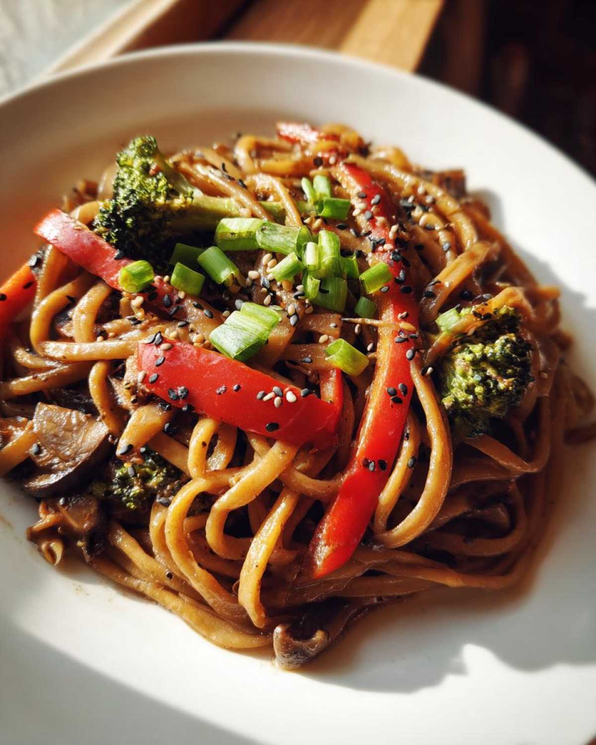 A close-up of a serving of Udon Stir-Fry with Sesame Vegetables, featuring thick noodles, red peppers, broccoli, and sesame seeds.